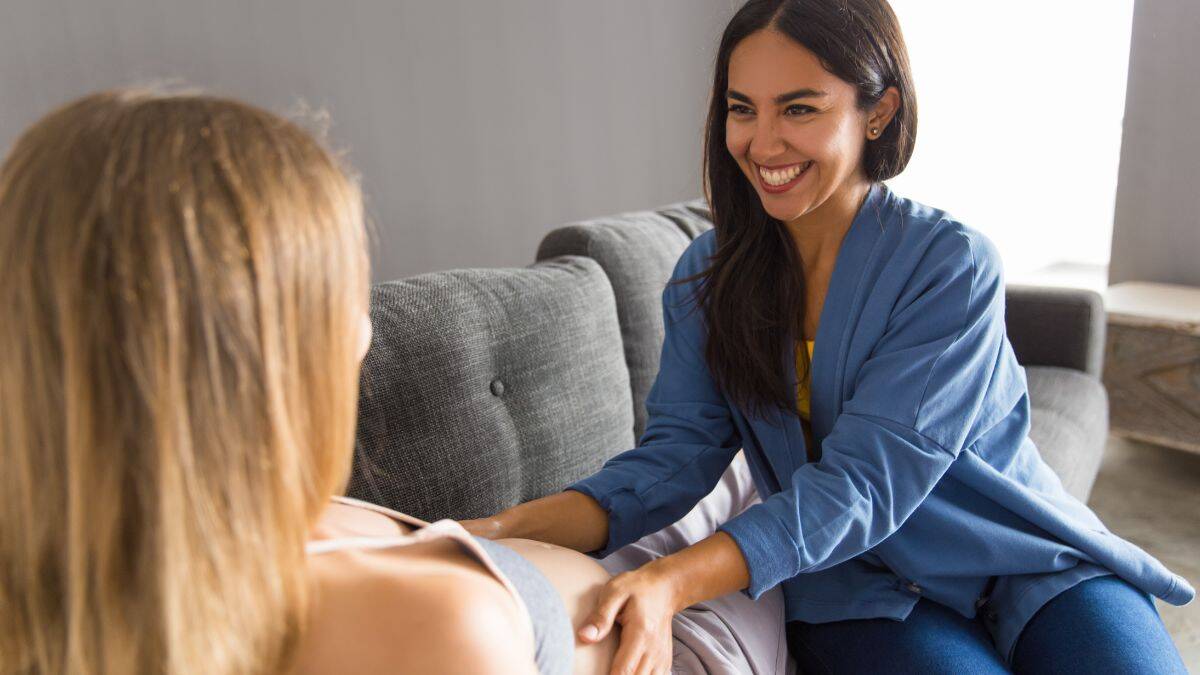 community midwife in plain clothes feeling the pregnant abdomen of a lady reclining on a couch in her home